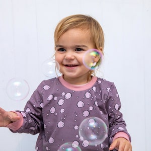 May include: A young child wearing a purple long-sleeved shirt with a pink bubble pattern is smiling and looking at bubbles floating in the air.