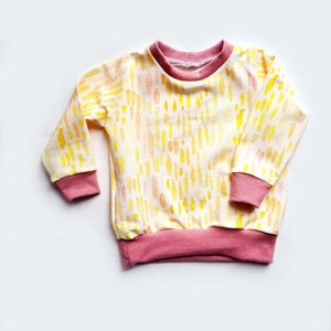 May include: A yellow and orange long-sleeved shirt with a pink trim. The shirt has a watercolor-like pattern of abstract shapes.