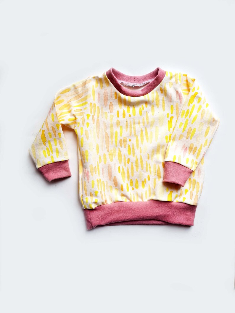 May include: A yellow and orange long-sleeved shirt with a pink trim. The shirt has a watercolor-like pattern of abstract shapes.