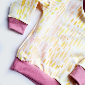 May include: A long-sleeved, white baby onesie with a yellow and orange watercolor pattern. The onesie has pink ribbed cuffs and a pink ribbed hem.
