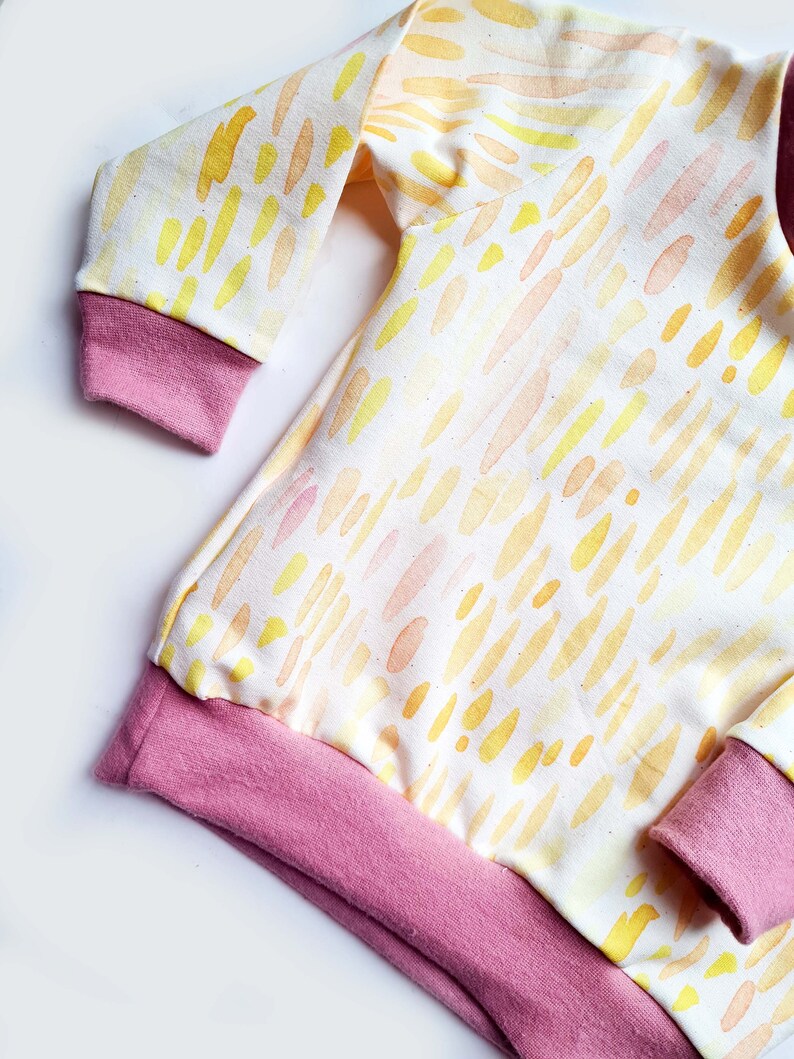 May include: A long-sleeved, white baby onesie with a yellow and orange watercolor pattern. The onesie has pink ribbed cuffs and a pink ribbed hem.