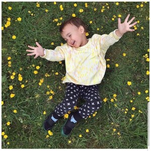 May include: A young child wearing a yellow and white tie-dye shirt and black pants with a white cross pattern. The child is lying on a bed of yellow flowers and smiling.