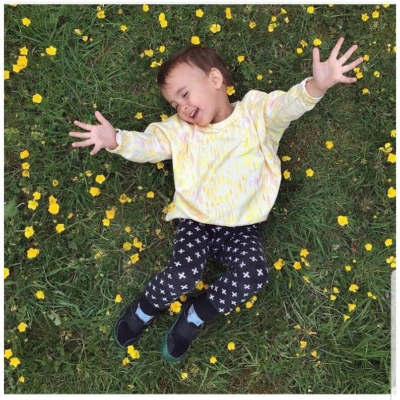 May include: A young child wearing a yellow and white tie-dye shirt and black pants with a white cross pattern. The child is lying on a bed of yellow flowers and smiling.