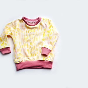 May include: A yellow and pink long-sleeved shirt with a watercolor-like pattern of yellow and orange dashes. The shirt has pink ribbed trim around the neck, cuffs, and hem.