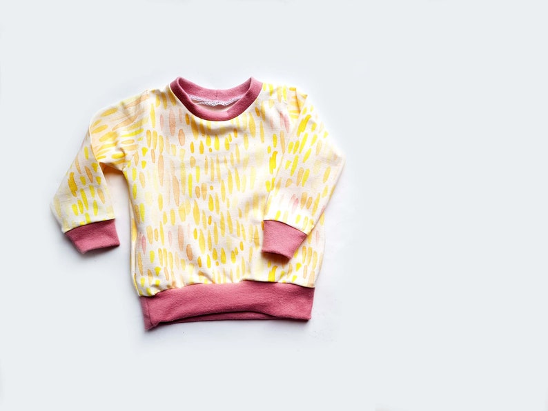 May include: A yellow and pink long-sleeved shirt with a watercolor-like pattern of yellow and orange dashes. The shirt has pink ribbed trim around the neck, cuffs, and hem.