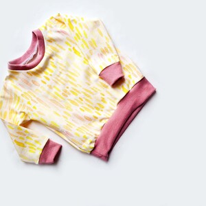 May include: A long-sleeved, yellow and pink baby sweater with a watercolor-like pattern. The sweater has a pink ribbed collar, cuffs, and hem.
