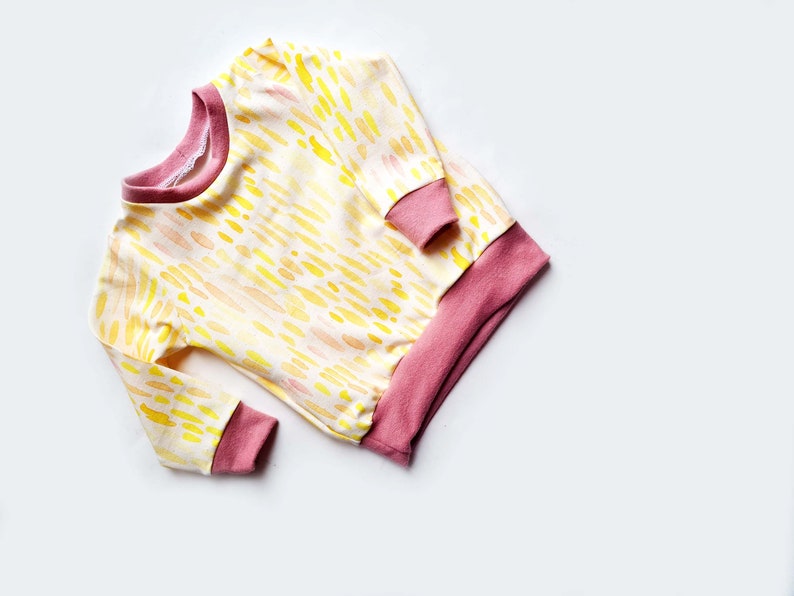 May include: A long-sleeved, yellow and pink baby sweater with a watercolor-like pattern. The sweater has a pink ribbed collar, cuffs, and hem.