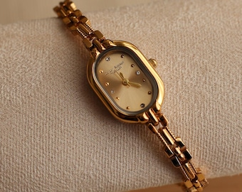 Puede incluir: Un reloj de pulsera dorado con una esfera ovalada y una correa de eslabones. La esfera del reloj es de color oro claro con pequeños detalles brillantes. La correa está hecha de pequeños eslabones rectangulares. El reloj se muestra sobre una superficie beige.