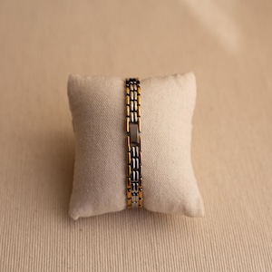 Pode incluir: Uma pulseira dourada e prateada com um design de elos retangulares. A pulseira &eacute; exibida em uma pequena almofada esbranqui&ccedil;ada contra um fundo neutro. Os elos da pulseira alternam entre ouro e prata.