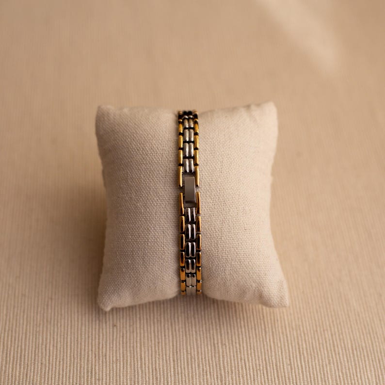 Pode incluir: Uma pulseira dourada e prateada com um design de elos retangulares. A pulseira &eacute; exibida em uma pequena almofada esbranqui&ccedil;ada contra um fundo neutro. Os elos da pulseira alternam entre ouro e prata.