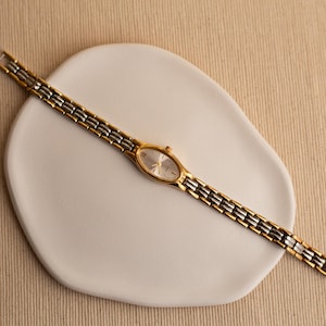 Pode incluir: Um rel&oacute;gio de pulso dourado e prateado com mostrador oval e pulseira de elos. O rel&oacute;gio &eacute; exibido em um prato branco de formato org&acirc;nico sobre um fundo neutro. O mostrador do rel&oacute;gio &eacute; prateado.