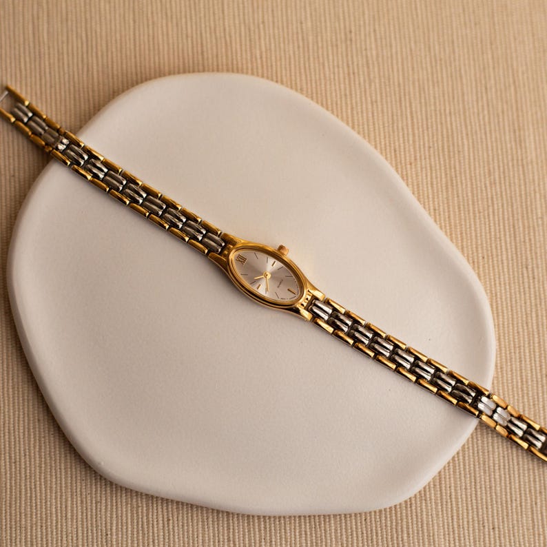 Pode incluir: Um rel&oacute;gio de pulso dourado e prateado com mostrador oval e pulseira de elos. O rel&oacute;gio &eacute; exibido em um prato branco de formato org&acirc;nico sobre um fundo neutro. O mostrador do rel&oacute;gio &eacute; prateado.