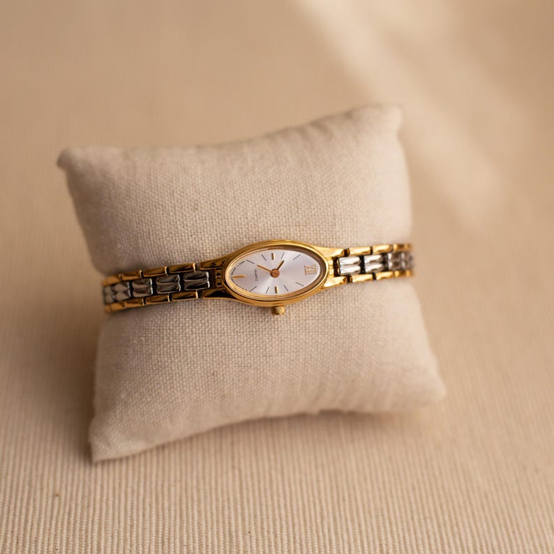 Pode incluir: Um rel&oacute;gio de pulso dourado e prateado com mostrador oval e pulseira dourada. O rel&oacute;gio &eacute; exibido em uma pequena almofada bege. O mostrador &eacute; branco com ponteiros e marcadores dourados. A pulseira &eacute; uma combina&ccedil;&atilde;o de elos dourados e prateados.
