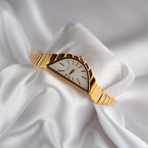 May include: A gold-toned wristwatch with a unique half-moon shaped face and a white dial. The watch features black hour markers and hands, and a ribbed gold band. The watch is displayed on a white, silky fabric.