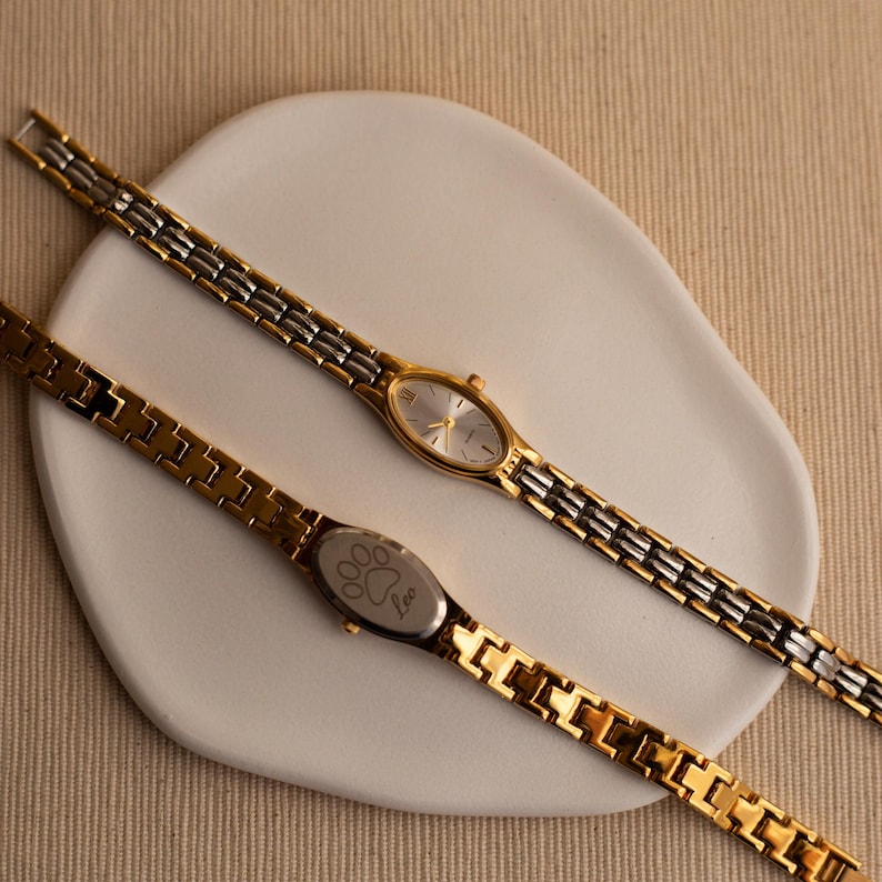 Pode incluir: Dois rel&oacute;gios de pulso dourados e prateados com mostradores ovais e pulseiras de elos repousam sobre um prato branco em forma org&acirc;nica. Um rel&oacute;gio tem um mostrador prateado, enquanto o outro tem um desenho de pata e a palavra "Leo" nas costas.