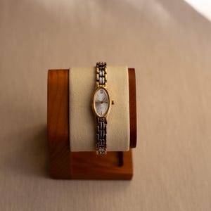Pode incluir: Um rel&oacute;gio de pulso dourado com um mostrador oval branco e uma pulseira bicolor dourada e prateada. O rel&oacute;gio &eacute; exibido em um suporte de madeira com uma almofada creme.