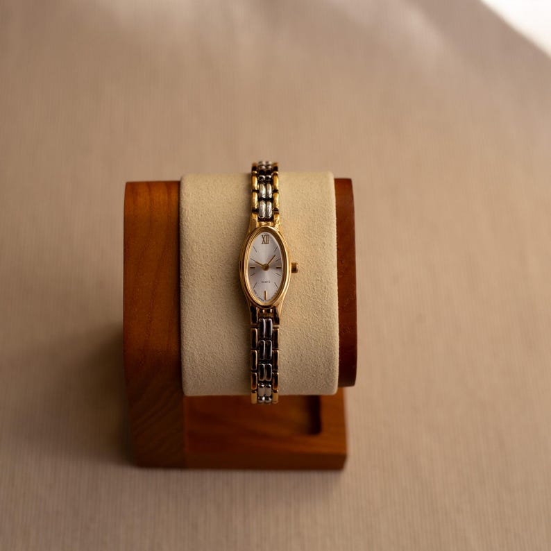 Pode incluir: Um rel&oacute;gio de pulso dourado com um mostrador oval branco e uma pulseira bicolor dourada e prateada. O rel&oacute;gio &eacute; exibido em um suporte de madeira com uma almofada creme.
