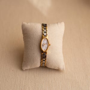 Pode incluir: Um rel&oacute;gio de pulso dourado e prateado com mostrador oval e pulseira de elos. O rel&oacute;gio &eacute; exibido em uma pequena almofada branca. O mostrador do rel&oacute;gio tem algarismos romanos na posi&ccedil;&atilde;o das 12 horas.