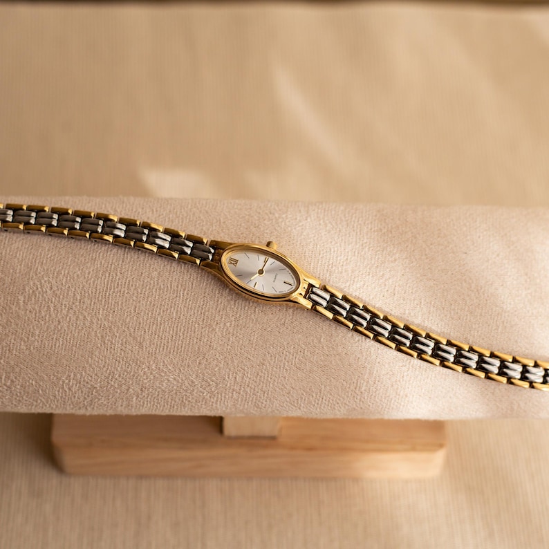 Pode incluir: Um rel&oacute;gio de pulso dourado e prateado com mostrador oval e pulseira de elos. O rel&oacute;gio tem um mostrador prateado com ponteiros dourados e algarismos romanos. A pulseira &eacute; uma combina&ccedil;&atilde;o de elos dourados e prateados.