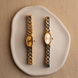 Pode incluir: Dois rel&oacute;gios de pulso ovais dourados com pulseiras de elos combinando s&atilde;o exibidos em um prato branco de formato org&acirc;nico. Um rel&oacute;gio tem mostrador dourado, o outro prateado. Os rel&oacute;gios est&atilde;o em um fundo neutro.