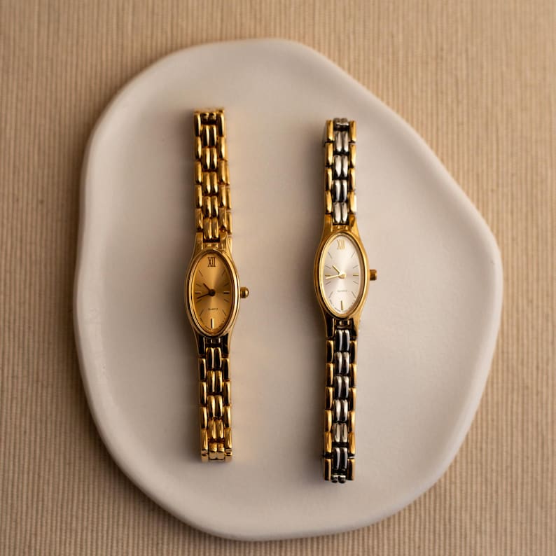 Pode incluir: Dois rel&oacute;gios de pulso ovais dourados com pulseiras de elos combinando s&atilde;o exibidos em um prato branco de formato org&acirc;nico. Um rel&oacute;gio tem mostrador dourado, o outro prateado. Os rel&oacute;gios est&atilde;o em um fundo neutro.