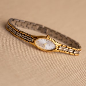 Pode incluir: Um rel&oacute;gio de pulso dourado e prateado com mostrador oval. O rel&oacute;gio tem uma moldura dourada e uma pulseira prateada. O mostrador &eacute; branco com ponteiros dourados e algarismos romanos. O rel&oacute;gio tem cerca de 18 cm de comprimento.