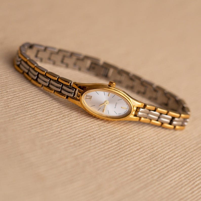 Pode incluir: Um rel&oacute;gio de pulso dourado e prateado com mostrador oval. O rel&oacute;gio tem uma moldura dourada e uma pulseira prateada. O mostrador &eacute; branco com ponteiros dourados e algarismos romanos. O rel&oacute;gio tem cerca de 18 cm de comprimento.