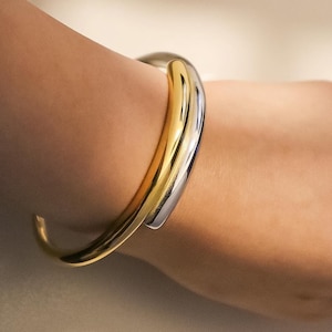 Op de afbeelding: Een goud- en zilverkleurige armband. De armband heeft een dik, afgerond ontwerp met een gouden band en een zilveren band die het goud overlapt. De armband wordt om een pols gedragen.