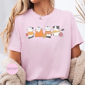 May include: A pale pink t-shirt with a graphic of four cartoon cats, each with a ball of yarn. The cats are wearing jumpers and scarves in various colours. The word "BLOSSOM" is printed on the shirt.