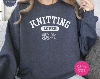 May include: A dark heather gray crewneck sweatshirt featuring the white text KNITTING LOYER in a collegiate font, accompanied by a graphic of a ball of yarn and two knitting needles. The garment has long sleeves and a ribbed neckline.
