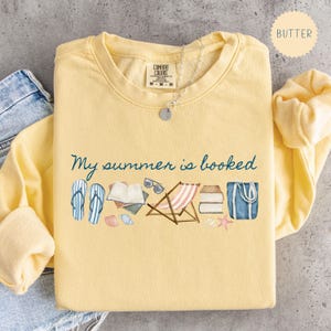 May include: A yellow Comfort Colors sweatshirt with the phrase "My summer is booked" in teal script. The design includes watercolor illustrations of flip-flops, books, sunglasses, a beach chair, and a tote bag. A silver necklace is draped on the shirt.