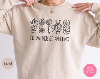 May include: A sand-colored crewneck sweatshirt featuring a black line-art graphic of a sock, a yarn ball with a heart, scissors, a yarn ball with needles, and a skein of yarn. Below the graphic, the text reads, I'D RATHER BE KNITTING.