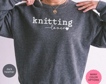 May include: A dark heather gray crewneck sweatshirt featuring the white text knitting lover with a small heart icon. The garment has long sleeves and a ribbed neckline. The design is centered on the chest area of the fabric.