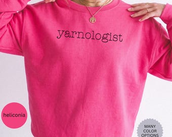 May include: A bright pink long-sleeve crewneck sweatshirt featuring the word "yarnologist" printed in black, lowercase, sans-serif lettering across the chest. The garment has a ribbed collar, cuffs, and hem, and is shown in a solid, vibrant color.