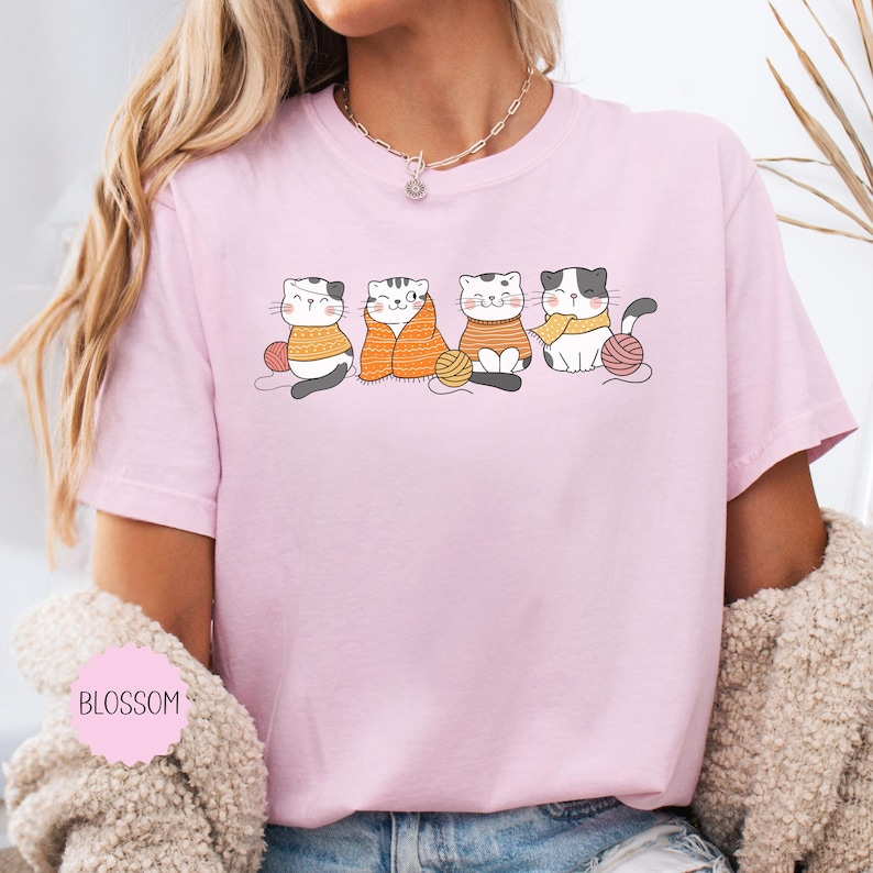 May include: A pale pink t-shirt with a graphic of four cartoon cats, each with a ball of yarn. The cats are wearing jumpers and scarves in various colours. The word "BLOSSOM" is printed on the shirt.