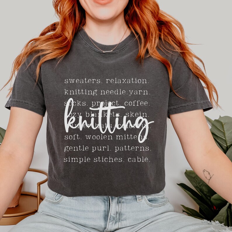 May include: Dark gray t-shirt with the word "knitting" in a cursive font. The shirt also features a list of knitting-related words in a white font, including "sweaters," "yarn," and "mittens."