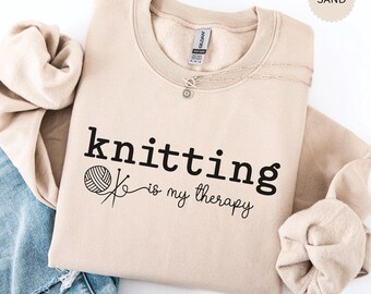 May include: A beige sweatshirt with the text "knitting is my therapy" in black. A small black and white illustration of a ball of yarn and knitting needles is to the left of the text.