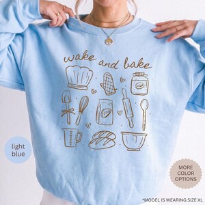 May include: A light blue crewneck sweatshirt featuring brown line art illustrations of baking tools including a chef hat, oven mitt, sugar jar, spatula, whisk, flour bag, rolling pin, spoon, measuring cup, bread loaves, and a mixing bowl with the text wake and bake.