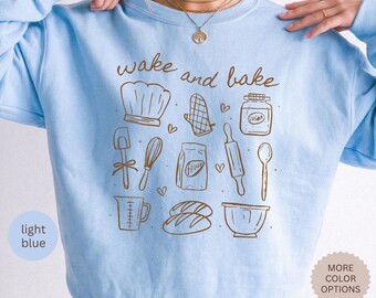 May include: A light blue crewneck sweatshirt featuring brown line art illustrations of baking tools including a chef hat, oven mitt, sugar jar, spatula, whisk, flour bag, rolling pin, spoon, measuring cup, bread loaves, and a mixing bowl with the text wake and bake.