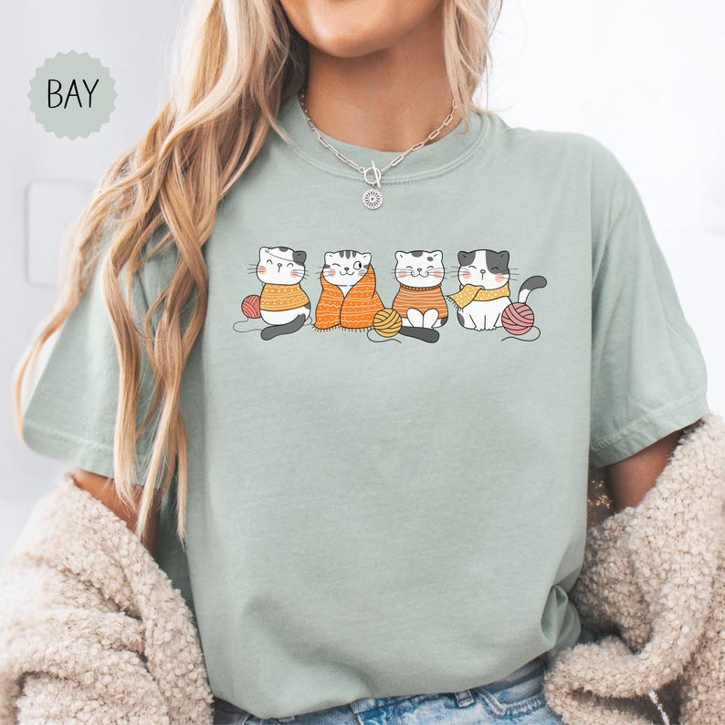 May include: A pale green t-shirt with a graphic of four cartoon cats, each with a ball of yarn. The cats are wearing sweaters and scarves in shades of orange and yellow. The word "BAY" is in a light green circle in the upper left.