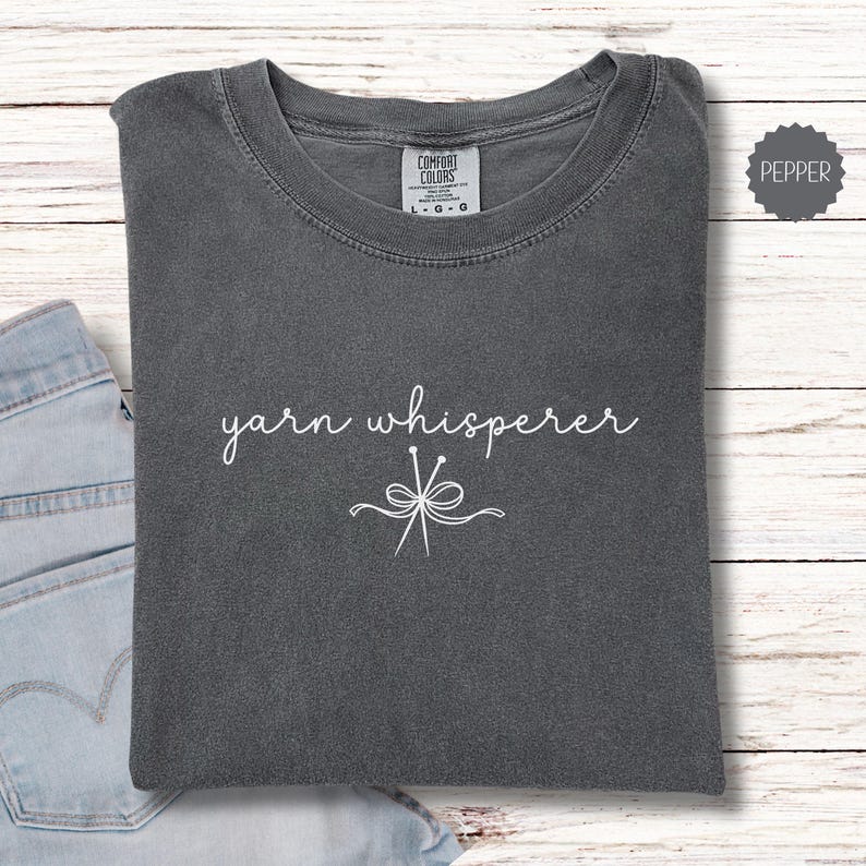 May include: A dark gray t-shirt with the words "yarn whisperer" in white script, along with a graphic of knitting needles and yarn. The shirt is folded and rests on a light blue denim surface.