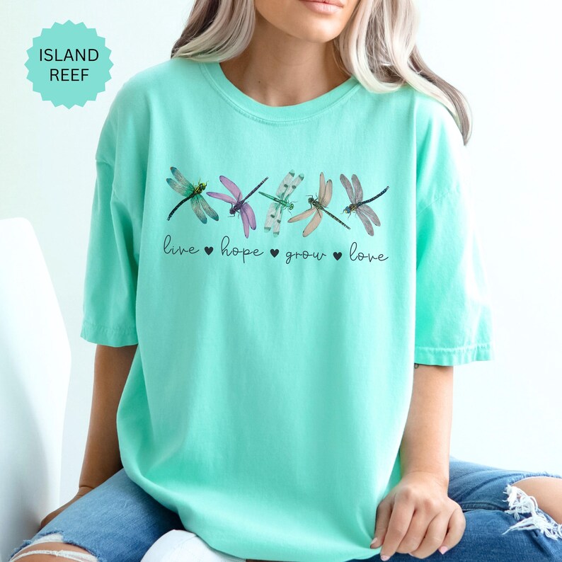以下が含まれることがあります： 明るいターコイズ色のTシャツ。カラフルなトンボのイラストが並んでいます。「live hope grow love」の文字が下にプリントされています。半袖でクルーネックです。