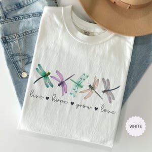 以下が含まれることがあります： カラフルなトンボのイラストが並んだ白いTシャツ。Tシャツには、トンボの下に「live hope grow love」という言葉がプリントされています。ベージュの帽子とブルージーンズも画像に写っています。
