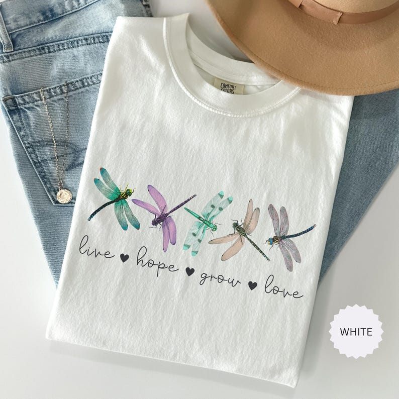 以下が含まれることがあります： カラフルなトンボのイラストが並んだ白いTシャツ。Tシャツには、トンボの下に「live hope grow love」という言葉がプリントされています。ベージュの帽子とブルージーンズも画像に写っています。