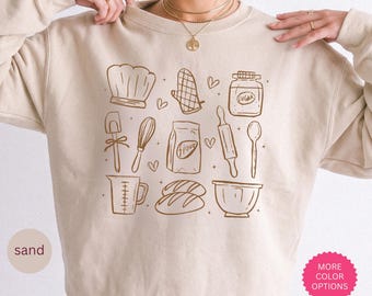 May include: A sand-colored crewneck sweatshirt featuring a brown line-art graphic of baking items including a chef hat, oven mitt, sugar jar, spatula, whisk, flour bag, rolling pin, spoon, measuring cup, bread loaves, and a mixing bowl.
