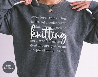 May include: A dark heather gray crewneck sweatshirt featuring white text that reads sweaters, relaxation, knitting needle, yarn, socks, project, coffee, cozy blankets, skein, knitting, soft, woolen mittens, gentle purl, patterns, simple stitches, cable.