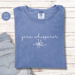 May include: A blue jean colored t-shirt with the words "yarn whisperer" in white script. Below the text is a white graphic of yarn needles and a bow. The shirt is folded on a light wood surface.