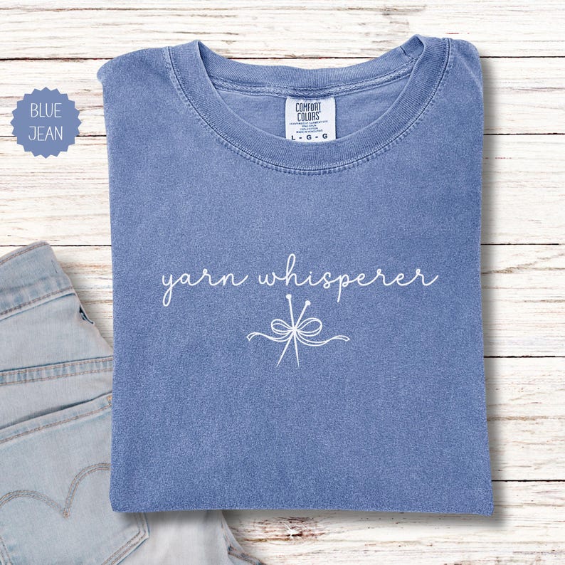 May include: A blue jean colored t-shirt with the words "yarn whisperer" in white script. Below the text is a white graphic of yarn needles and a bow. The shirt is folded on a light wood surface.