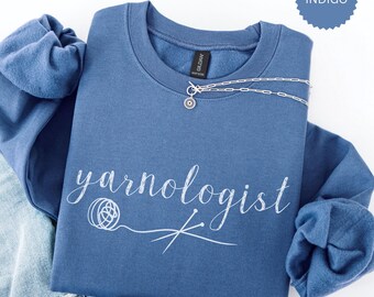 May include: Indigo blue sweatshirt with the text "yarnologist" in white cursive lettering. The text is accompanied by a white outline of a ball of yarn and knitting needles.