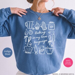 May include: An indigo blue crewneck sweatshirt featuring a white line-art graphic of baking tools and the text "Baking is my love language." The design includes illustrations of a measuring cup, cupcake, oven mitt, sugar jar, spatula, whisk, rolling pin, chef hat, piping bag, flour bag, and mixing bowl.